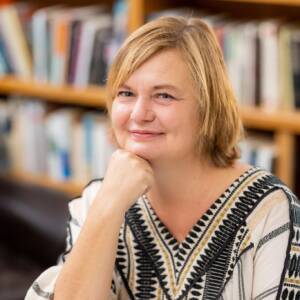 A woman with short blonde hair is sitting indoors with her chin resting on her hand. She is wearing a patterned blouse and smiling slightly. The background features a blurred bookshelf filled with books.
