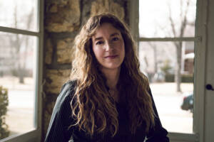 A woman with long curly hair and a relaxed smile stands indoors by a window, allowing natural light to illuminate her. She is wearing a dark, striped shirt and is positioned against a backdrop of an interior wall with large stones. Sparse trees and buildings are visible outside.