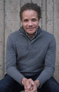 A person with shoulder-length dreadlocks, wearing a gray sweater and dark pants, sits against a stone wall. They are smiling slightly and have their hands clasped in front of them.