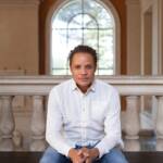 A man wearing a white shirt and blue jeans sits on a bench indoors in front of a large arched window with a bright outdoor view in the background. He has his hands clasped and looks directly at the camera. The setting appears to be a historical or classical building.