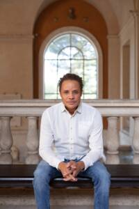 A man wearing a white shirt and blue jeans sits on a bench indoors in front of a large arched window with a bright outdoor view in the background. He has his hands clasped and looks directly at the camera. The setting appears to be a historical or classical building.