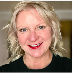 A person with short, wavy blonde hair smiles at the camera, wearing a dark top and red lipstick. The background is softly blurred.