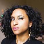 Person with curly black hair and a thoughtful expression, wearing a dark top and dangling earrings. The background is softly blurred with a hint of colorful lights.