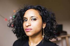 Person with curly black hair and a thoughtful expression, wearing a dark top and dangling earrings. The background is softly blurred with a hint of colorful lights.