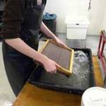 A person is using a screen to make paper by hand. They are dipping the screen into a water-filled tray, with a white sink in the background. The setting appears to be a workshop or studio.
