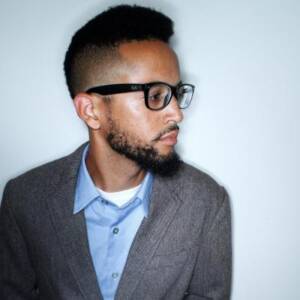 A man with short, curly hair and a beard is wearing black-rimmed glasses, a blue collared shirt, and a gray blazer. He is looking to his right against a plain white background.