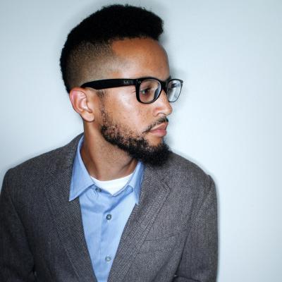 A man with short, curly hair and a beard is wearing black-rimmed glasses, a blue collared shirt, and a gray blazer. He is looking to his right against a plain white background.