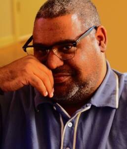A person with short hair and glasses is wearing a blue collared shirt and gazing towards the camera with a slight smile, resting their chin on their hand. The background is softly blurred, with warm lighting.