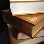 A close-up of three stacked books with ornate golden designs on the covers. The books are positioned at different angles, and the pages are slightly yellowed, giving them an antique appearance.