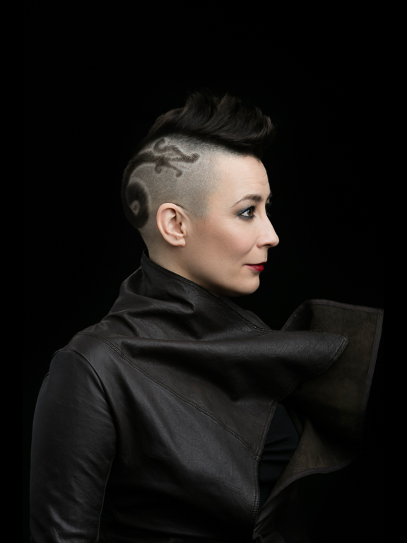 A person with a mohawk hairstyle and a shaved design on the side of their head faces left. They're wearing a dark, structured leather jacket against a black background.