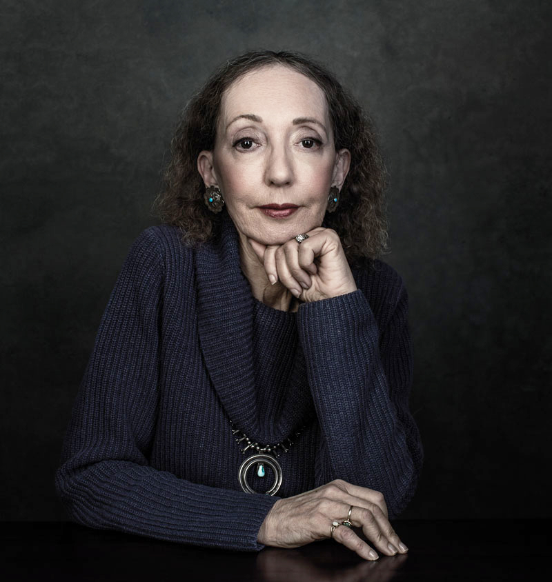 Person with curly hair and a thoughtful expression, resting chin on hand. Wearing a dark ribbed sweater, large pendant necklace, and earrings. Plain dark background.