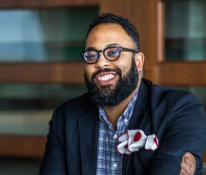A man with glasses and a beard smiles while wearing a dark jacket and a checkered shirt. A red and white pocket square is visible in his jacket. The background features wooden shelves and a blurred room.