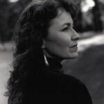A woman with long curly hair is captured in black and white, gazing to her right. She is wearing earrings and a dark top. The background is blurred, showing a hint of trees and a path, giving an outdoor setting.