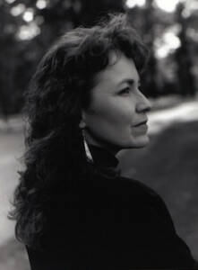 A woman with long curly hair is captured in black and white, gazing to her right. She is wearing earrings and a dark top. The background is blurred, showing a hint of trees and a path, giving an outdoor setting.