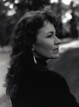 A woman with long curly hair is captured in black and white, gazing to her right. She is wearing earrings and a dark top. The background is blurred, showing a hint of trees and a path, giving an outdoor setting.