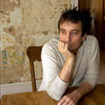 Man with dark hair and wearing a light gray shirt sits at a wooden table, resting his chin on his fist, looking thoughtfully to the side. The background features a textured, rustic wall.