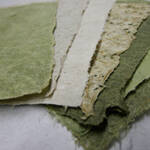 A close-up of various pieces of textured, handmade paper in shades of green and off-white. The papers are layered, revealing rough and uneven edges, highlighting their artisanal quality.