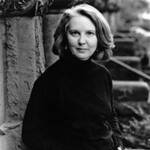 Black and white portrait of a person with shoulder-length hair, wearing a dark turtleneck. They are standing outdoors, leaning against a stone wall, with stairs visible in the background.