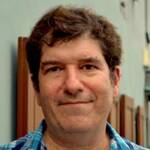 A person with short, curly brown hair and a slight smile stands in front of a building with light blue walls and brown shutters. They are wearing a blue and green plaid shirt. The background is slightly out of focus.