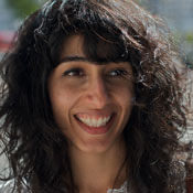 A woman with curly dark hair smiles warmly. She is outside with a blurred background of trees and buildings, wearing a white top. The lighting suggests it's a sunny day.