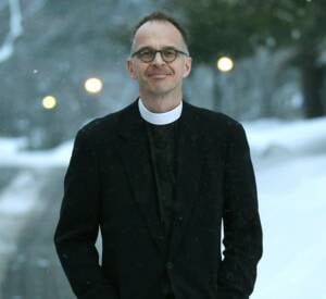 A person wearing glasses and a clerical collar stands outdoors in a snowy landscape. They are dressed in a dark suit and smiling softly. Blurred lights and snow-covered trees are visible in the background.