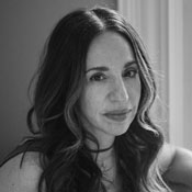 Black and white portrait of a woman with long, wavy hair. She gazes softly at the camera, with a subtle, relaxed expression. The background is a simple, light-colored wall.