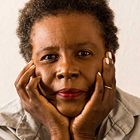A person with short curly hair rests their chin on their hands, looking directly at the camera. They wear a light-colored shirt and have a calm expression. Their nails are painted white. The background is plain and light-colored.