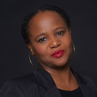 A person with braided hair is wearing hoop earrings, a dark blazer, and red lipstick, set against a dark background.