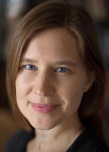 A person with shoulder-length brown hair smiles softly, looking directly at the camera. They have blue eyes and are wearing a dark top. The background is blurred.
