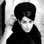 Black and white photo of a person in a dark coat and large textured hat, looking up at the camera. The background is blurred, highlighting the subject's expression and attire.