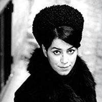 Black and white photo of a person in a dark coat and large textured hat, looking up at the camera. The background is blurred, highlighting the subject's expression and attire.