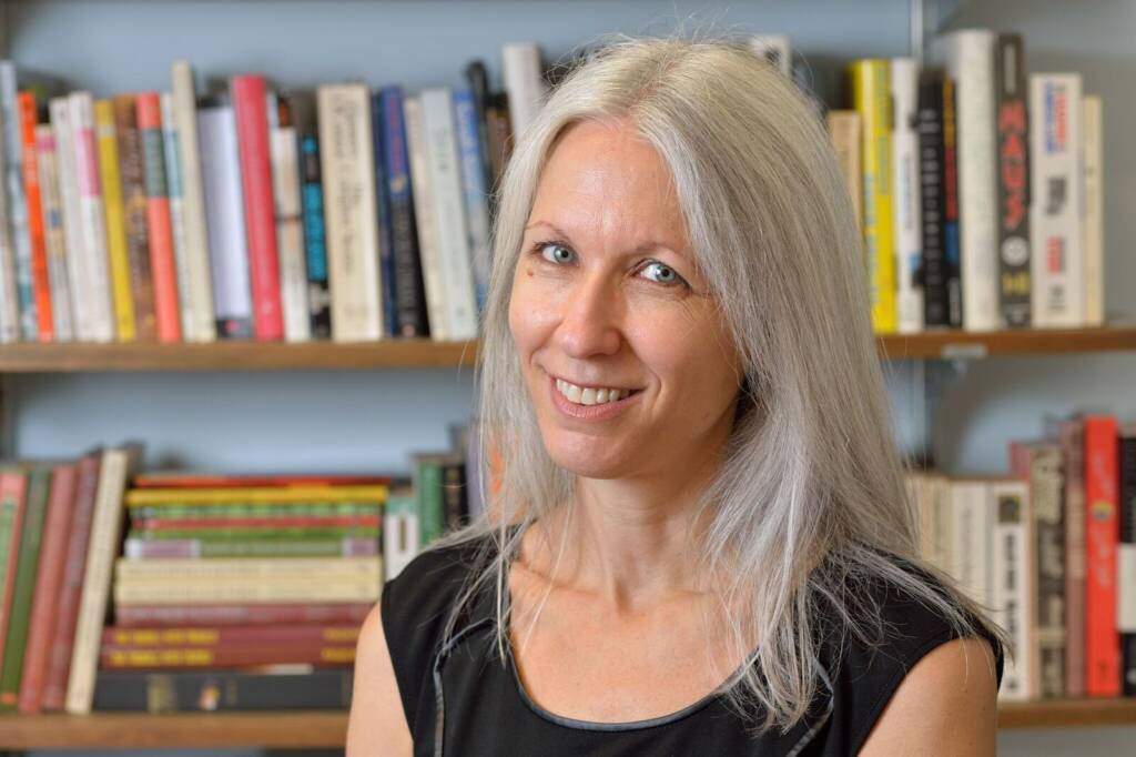 A woman with long gray hair and a black sleeveless top smiles in front of bookshelves filled with colorful books.
