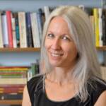 A woman with long gray hair and a black sleeveless top smiles in front of bookshelves filled with colorful books.