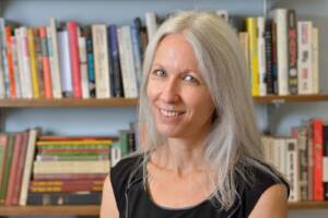 A woman with long gray hair and a black sleeveless top smiles in front of bookshelves filled with colorful books.