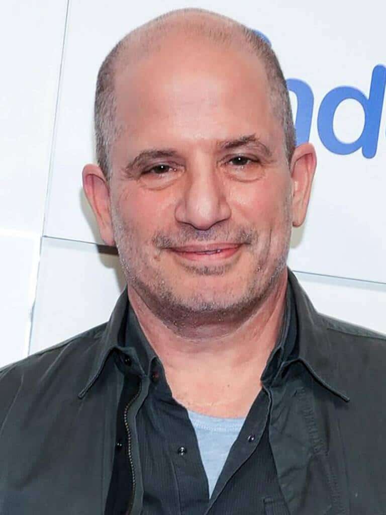 A middle-aged man with a shaved head and slight stubble smiles at the camera. He is wearing a gray shirt with a dark jacket and is standing in front of a white background with blue text.