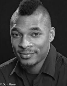 Black and white portrait of a man with a short mohawk hairstyle, trimmed beard, and mustache, wearing a dark collared shirt, looking at the camera with a slight smile. © Don Usner.