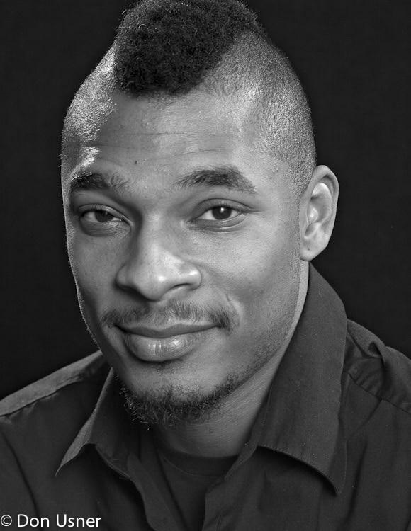 Black and white portrait of a man with a short mohawk hairstyle, trimmed beard, and mustache, wearing a dark collared shirt, looking at the camera with a slight smile. © Don Usner.