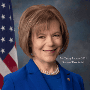 A woman with short brown hair, wearing a blue blazer and pearl necklace, smiles in front of the U.S. flag. The text "McCarthy Lecture 2021 Senator Tina Smith" is on the right side of the image.