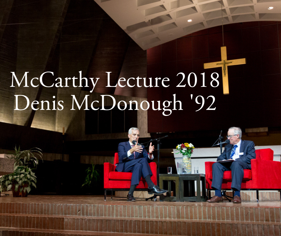 Two speakers seated on a stage in front of a large cross in a church-like setting. The slide behind them reads "McCarthy Lecture 2018 Denis McDonough '92." Both men are engaged in discussion, surrounded by greenery and colorful flowers on a table.