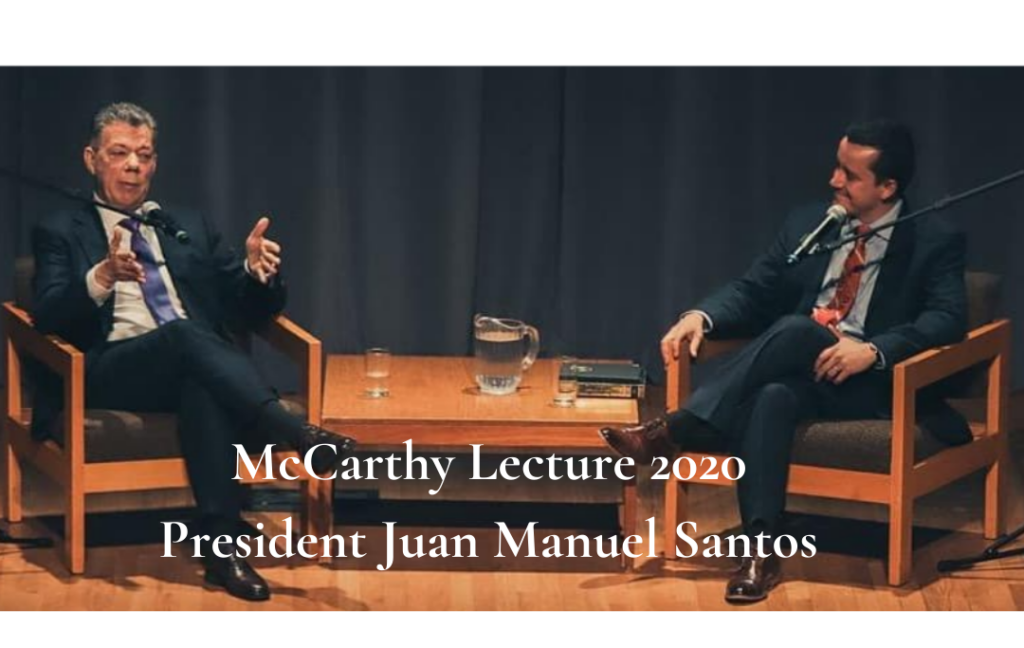 Two men are seated on a stage in armchairs, engaged in conversation. The one on the left is speaking with hand gestures. A table with a water pitcher, glasses, and books is between them. Text overlay reads: "McCarthy Lecture 2020 President Juan Manuel Santos.