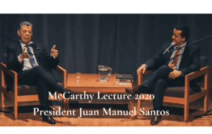 Two men are seated on a stage in armchairs, engaged in conversation. The one on the left is speaking with hand gestures. A table with a water pitcher, glasses, and books is between them. Text overlay reads: "McCarthy Lecture 2020 President Juan Manuel Santos.