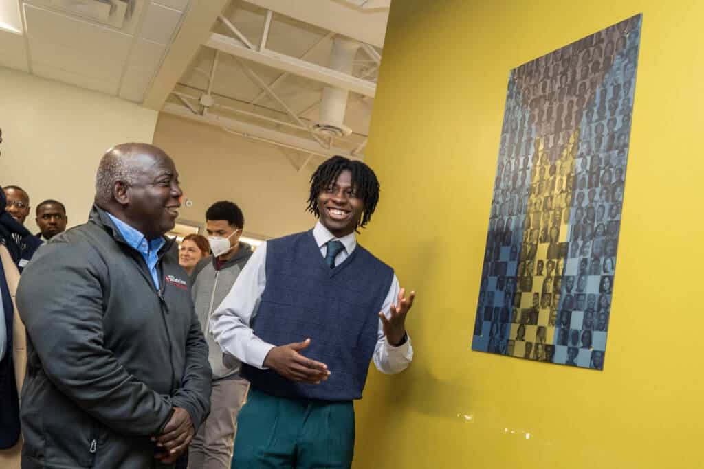 A smiling young man in a vest gestures toward artwork on a yellow wall while an older man beside him and others in the background watch and smile in a bright indoor setting.