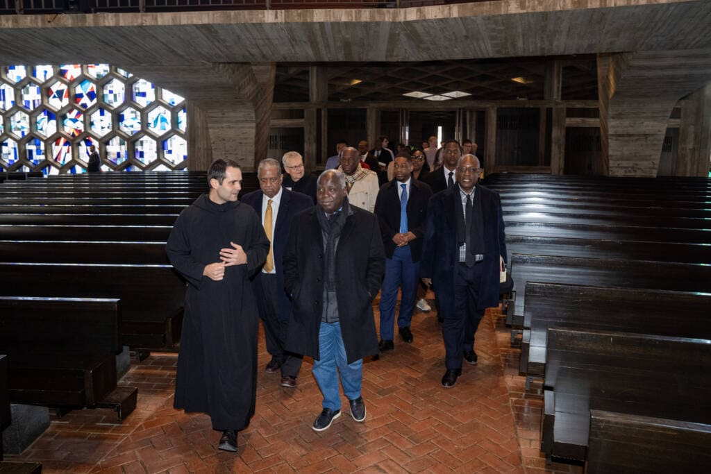 A group of men, including a priest in black robes, walk through the aisle of a church with wooden pews and colorful stained glass windows. The group appears to be in conversation during a tour or visit.