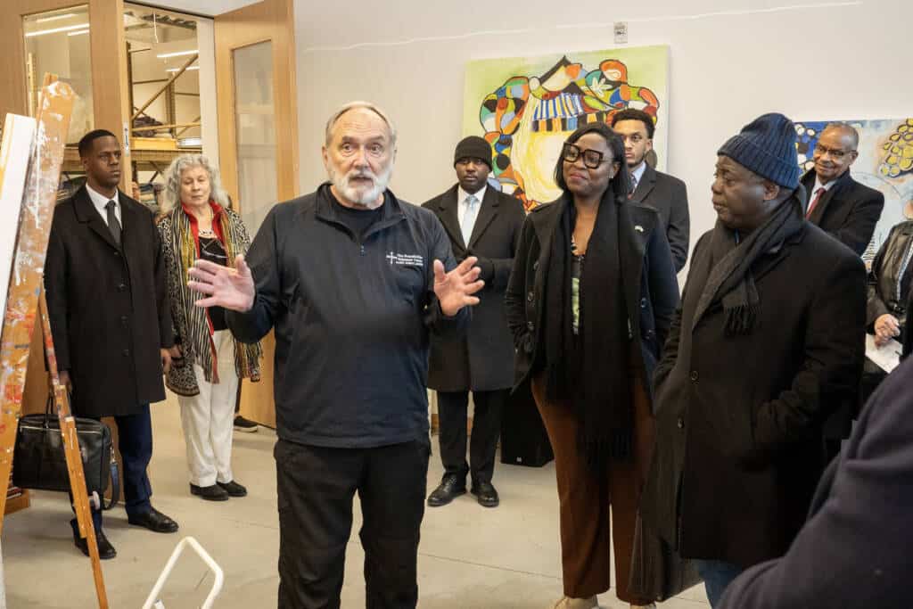 A man with gray hair and a beard speaks to a group of people in an art studio. The group listens attentively, with paintings and colorful artwork visible in the background.