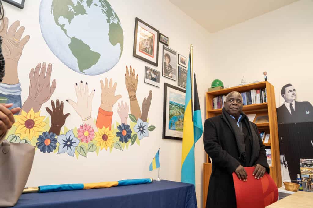 A man in a dark coat stands beside a desk in an office with a mural of diverse raised hands and a globe on the wall, a Bahamas flag, and shelves with books and framed photos.
