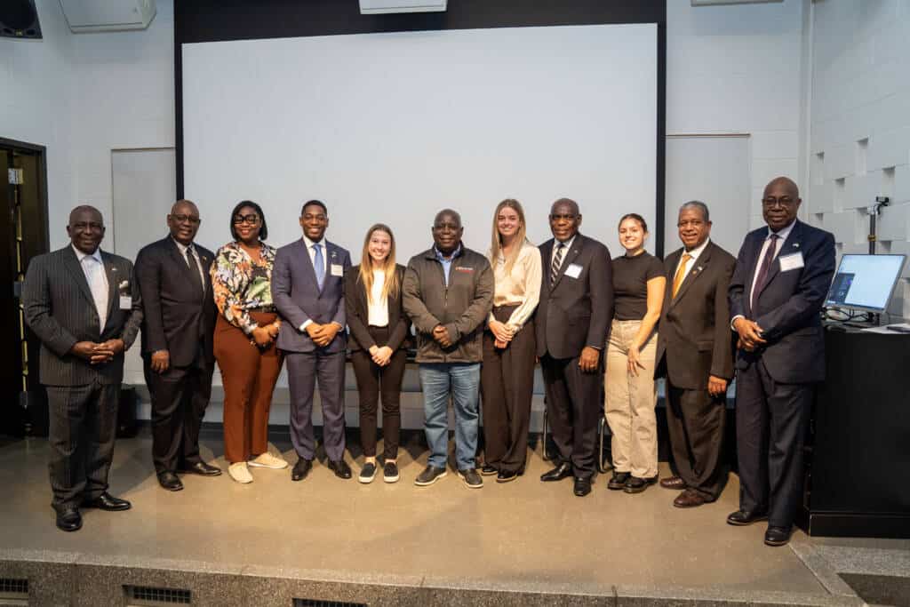 A group of twelve people, including men in suits and women in business or casual attire, stand side by side and smile for a group photo in front of a large projector screen in a lecture room.