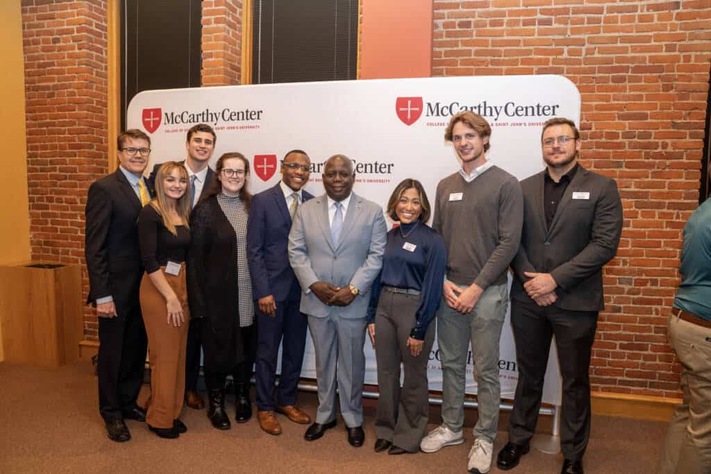 A group of people, dressed in business and smart casual attire, stand together smiling in front of a McCarthy Center banner in a room with brick walls and large windows.