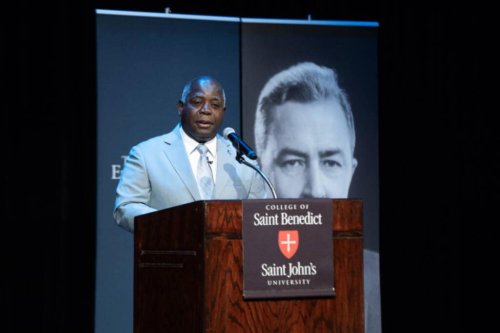 A man in a light blue suit speaks at a podium with a microphone. The podium displays the College of Saint Benedict and Saint John’s University logos. A large black-and-white portrait is visible in the background.