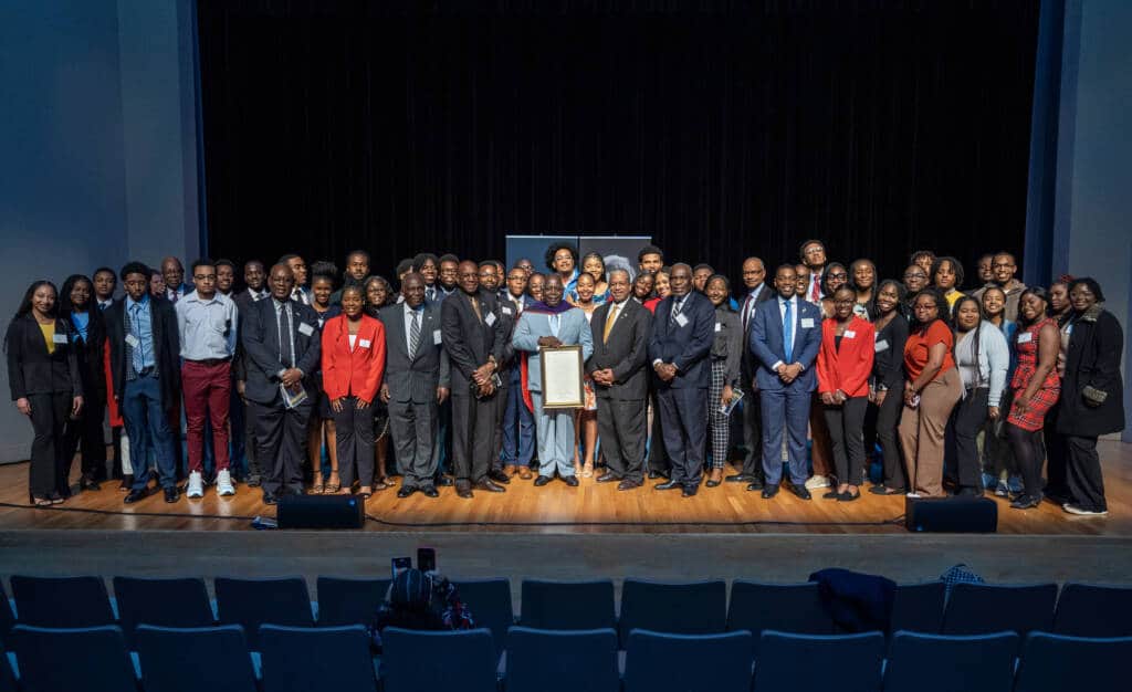 A large group of people in formal attire pose together on a stage in front of a black curtain, with some holding a framed document; there are empty seats visible in the foreground.