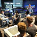 A group of people is seated in a modern office setting, engaged in a discussion. A man stands, speaking to the group, while others sit facing him. Panasonic branding is visible in the background.
